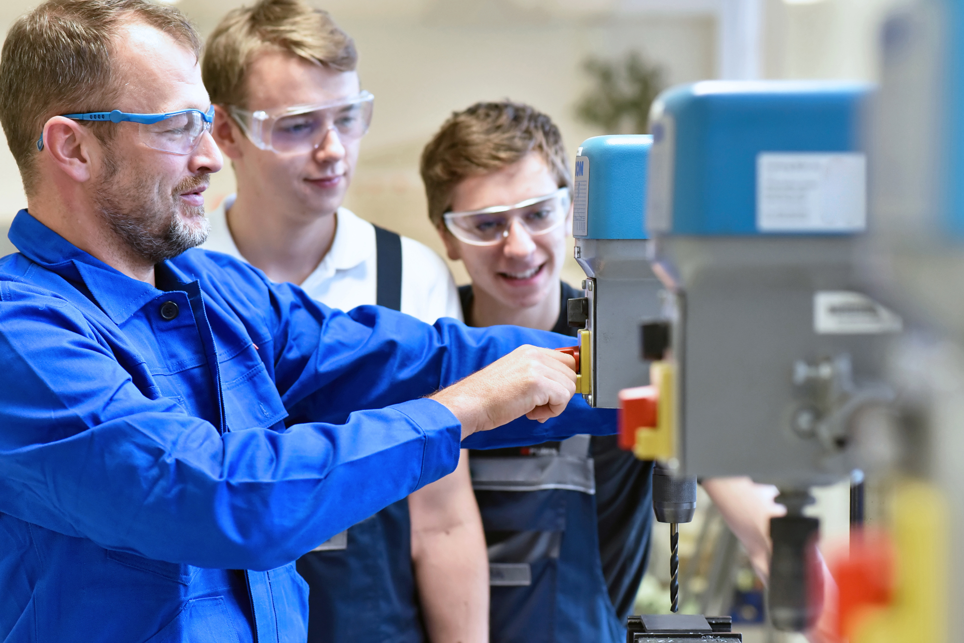 Ausbilder zeigt zwei Auszubildenden den Umgang mit einer Bohrmaschine in einer Lehrwerkstatt.