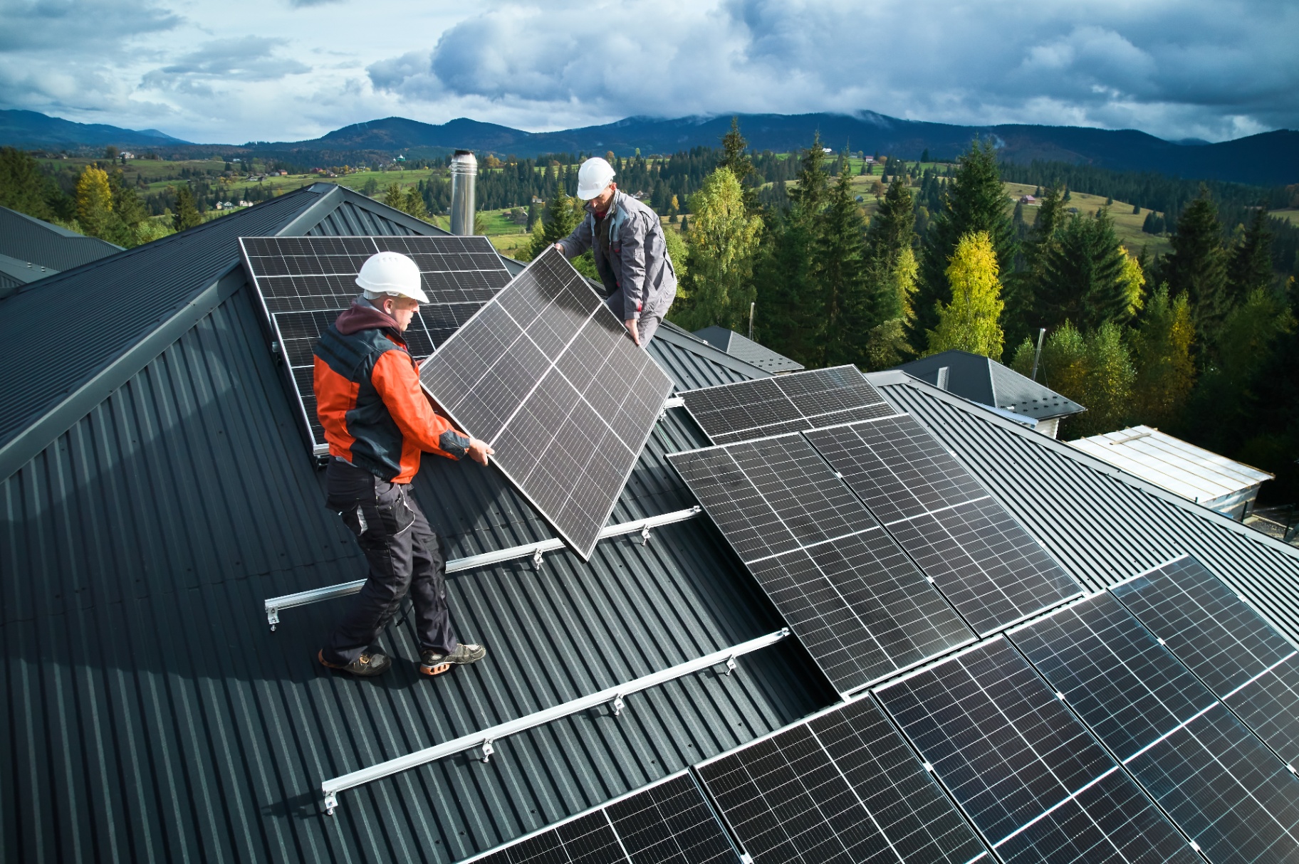 Zwei Handwerker montieren Solarmodule auf einem Schrägdach in bergiger Umgebung.